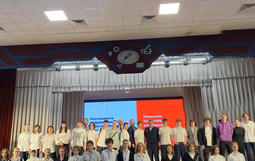 Группа людей стоит на сцене в актовом зале. На сцене висят шторы и экран с текстом.