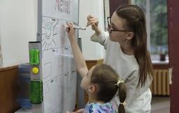 На изображении две девушки: одна взрослая, другая маленькая. Они рисуют на белой доске. 