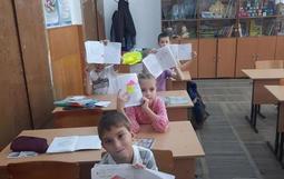 В классе сидят дети за партами, держат рисунки. На стенах учебные плакаты и глобус. На полках книги и учебные материалы.