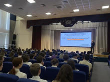 В помещении с рядами синих кресел проходит лекция. На сцене человек стоит перед экраном с текстом