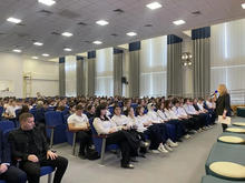 Фото с мероприятия.В большом светлом зале сидят школьники в форме, слушая выступающего человека с микрофоном