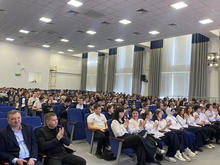 Фото с мероприятия. На изображении большой зал с рядами стульев, заполненными людьми. Люди аплодируют.