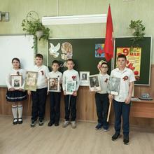 Шесть детей в белых рубашках и тёмных брюках стоят в классе. У каждого в руках фото. На доске плакат ко Дню Победы.