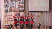 На сцене дети в красных юбках и чёрных костюмах с красными шляпами. На заднем плане баннер с военной тематикой и надписью «Помним, гордимся и чтим!».