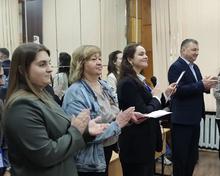 На изображении группа людей стоит в помещении, аплодирует. Люди разного возраста, одеты в разную одежду, некоторые в деловом стиле.