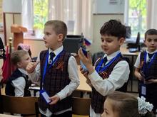 На изображении несколько детей в школьной форме с жилетами и лентами. Они стоят в классе, держа в руках бейджи.