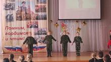 На сцене четыре ребёнка в военной форме. На заднем плане баннер с надписью «Помним, гордимся и чтим!». Также видны звёзды и экран.