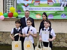 На изображении трое детей и двое взрослых. Дети держат сумки и улыбаются. На заднем плане баннер с надписью «Ключевые принципы ЮИД». Также видны воздушные шары.