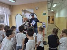 На изображении группа детей в белых футболках, стоящих полукругом. Перед ними мужчина и кукла в детском кресле. Помещение светлое, с жёлтыми стенами и окнами.