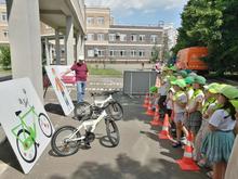 На изображении дети в зелёных шляпах и белых рубашках стоят в ряд возле оранжевых конусов. Рядом велосипеды и плакаты с рисунками.