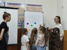 На изображении группа детей и двое взрослых в помещении. На стенах плакаты с дорожными знаками. На доске разноцветные круги.