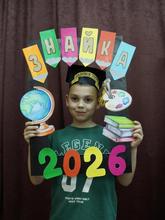 На изображении мальчик  держит картонную рамку с надписью «ЗНАЙКА 2026». На рамке также изображены глобус, палитра и книги.