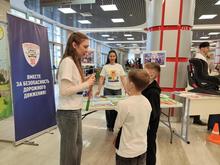 На изображении люди в белых футболках, дети и взрослые, стоят вокруг стола с материалами. Помещение светлое, с колонной и баннером с надписью :" Вместе за безопасность дорожного движения."