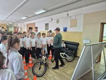 В помещении группа детей в белых футболках слушает мужчину в зелёной кофте. На полу оранжевые конусы и велосипеды. Рядом доска с информацией.