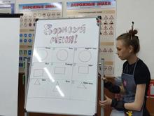 На изображении девушка стоит у доски. На доске задание «Дорисуй меня!». Вокруг доски висят плакаты с дорожными знаками.