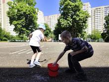 На изображении двое детей: один в белой футболке и голубой шляпе, другой в тёмной футболке. Они играют на асфальтовой площадке возле красного ведра.