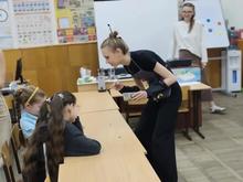 В классе девушка в чёрном наклоняется к двум девочкам за партой. На заднем плане стоит другая девушка у доски. Класс украшен плакатами и учебными материалами.