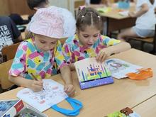 На изображении две девочки в разноцветных футболках. Одна из них рисует, другая держит книгу с картинками. Они сидят за столом в классе.