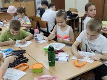 На изображении дети сидят за столами, рисуют и раскрашивают. На столах лежат фломастеры, цветные карандаши, бумага и вода.