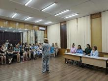 В комнате с деревянными стенами и зеркалами сидят родители детей из объединения "Знайка", слушают педагога, стоящего перед ними. Она рассказывает о начале учебного года и об организационных моментах. На столах лежат бумаги и вода.