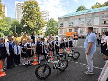 На изображении группа детей в школьной форме стоит на улице возле двух велосипедов. Рядом педагог объясняет устройство велосипеда