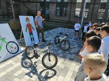 На изображении группа детей стоит полукругом, слушая человека с плакатом. На земле два велосипеда, рядом плакат с изображением ребёнка в шлеме.