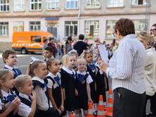 На изображении группа детей в школьной форме, стоящих на улице. Рядом с ними педагог, который объясняет задание. На заднем плане здание и оранжевый фургон "Лаборатория безопасности".