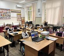 В школьном классе проходит занятие, посвящённое Дню народного единства: ученики в форме сидят за партами, слушают рассказ об истории праздника. На столе учителя — ноутбук с тематическим слайдом, проектор; на стенах — патриотические плакаты (в т. ч. с надписью «Никто не забыт, ничто не забыто»)