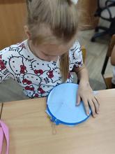 На изображении девочка с длинными светлыми волосами, одетая в футболку с рисунком. Она сидит за столом и вышивает орнамент