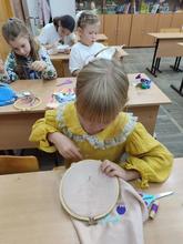 На изображении дети занимаются рукоделием в классе. Девочка в жёлтом платье шьёт на пяльцах орнамент в виде цветов