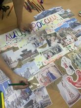 На изображении стол с фотографиями, рисунками и надписями о России. Видны руки ребенка, который что-то пишет зелёным маркером.