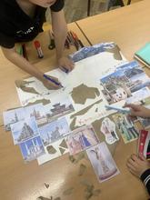 На изображении стол с плакатом и фотографиями достопримечательностей. Двое детей работают с плакатом, используя маркеры и клей.